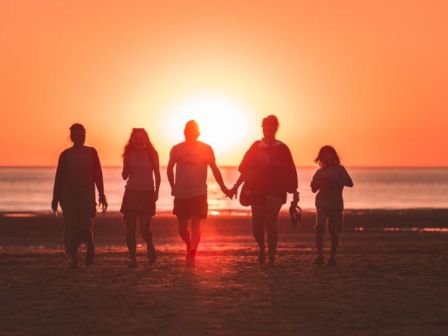 A group of five people is walking on the beach at sunset, silhouetted against the orange sky while holding hands.