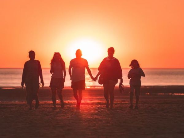 A group of five people is walking on the beach at sunset, silhouetted against the orange sky while holding hands.