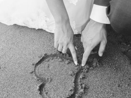 A bride and groom draw a heart in the sand with their fingers, celebrating their wedding in a romantic beach setting, showcasing their love.