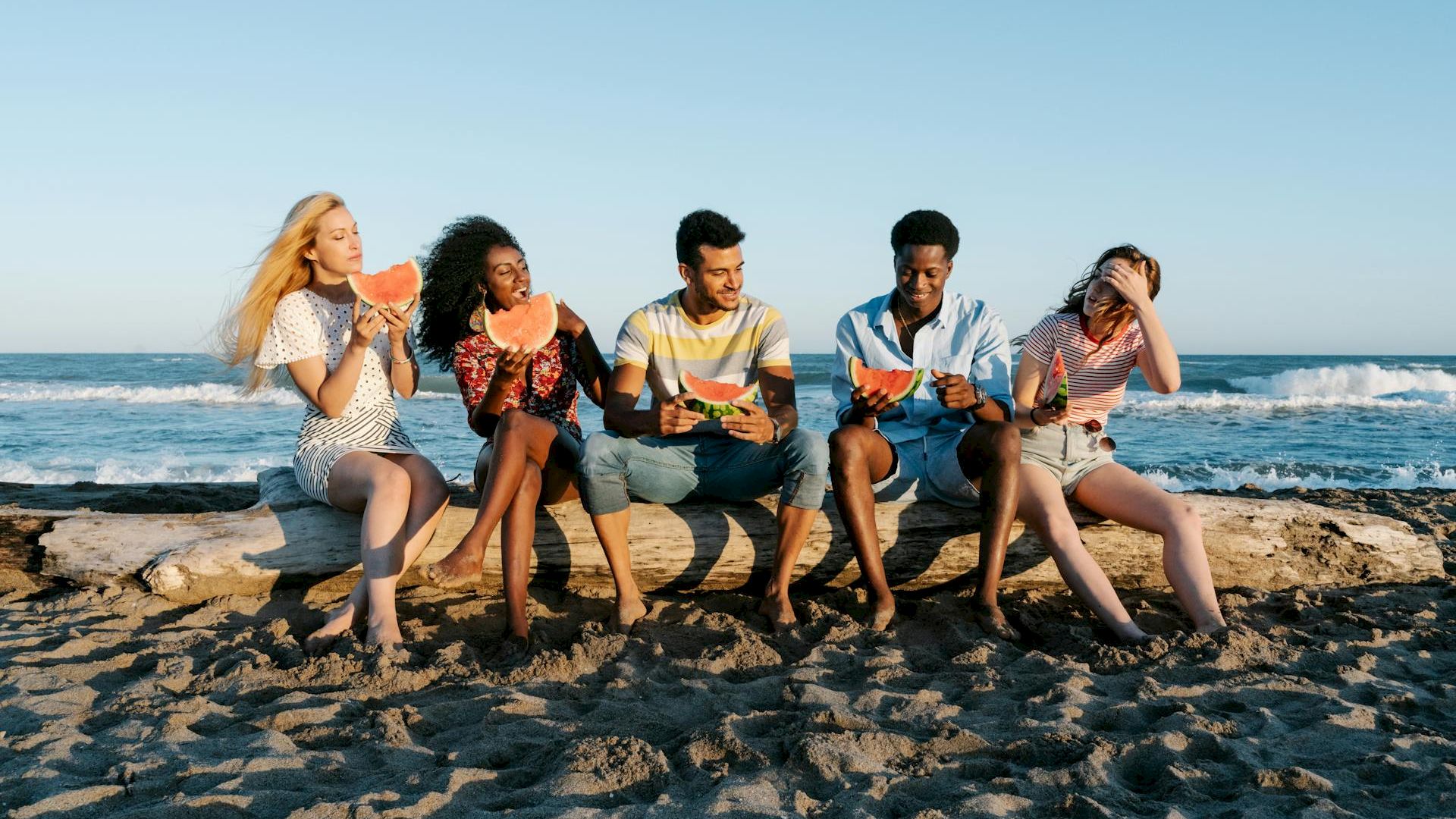 Five people sitting on a beach log, enjoying slices of watermelon by the ocean.