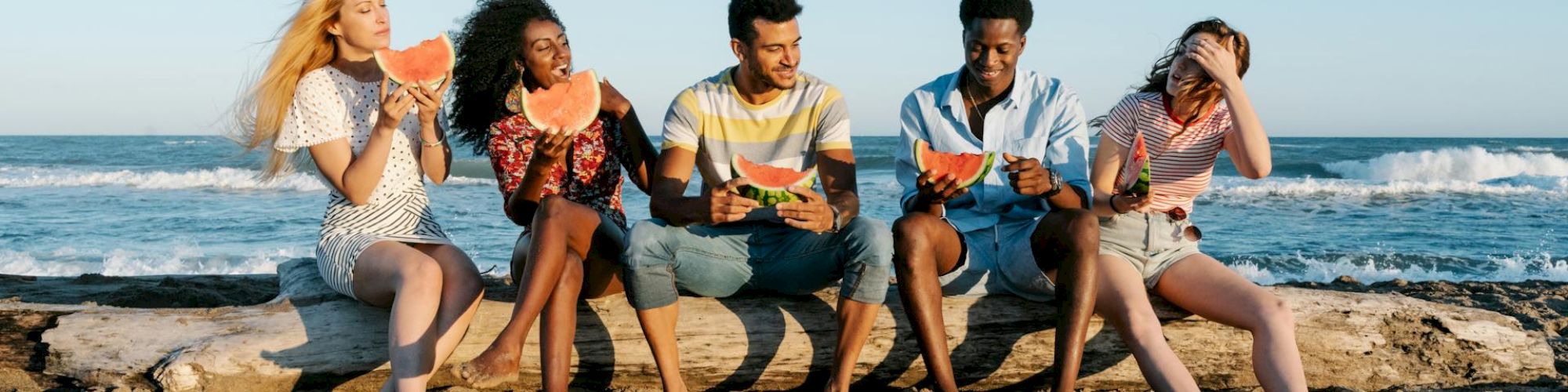 Five people sitting on a beach log, enjoying slices of watermelon by the ocean.