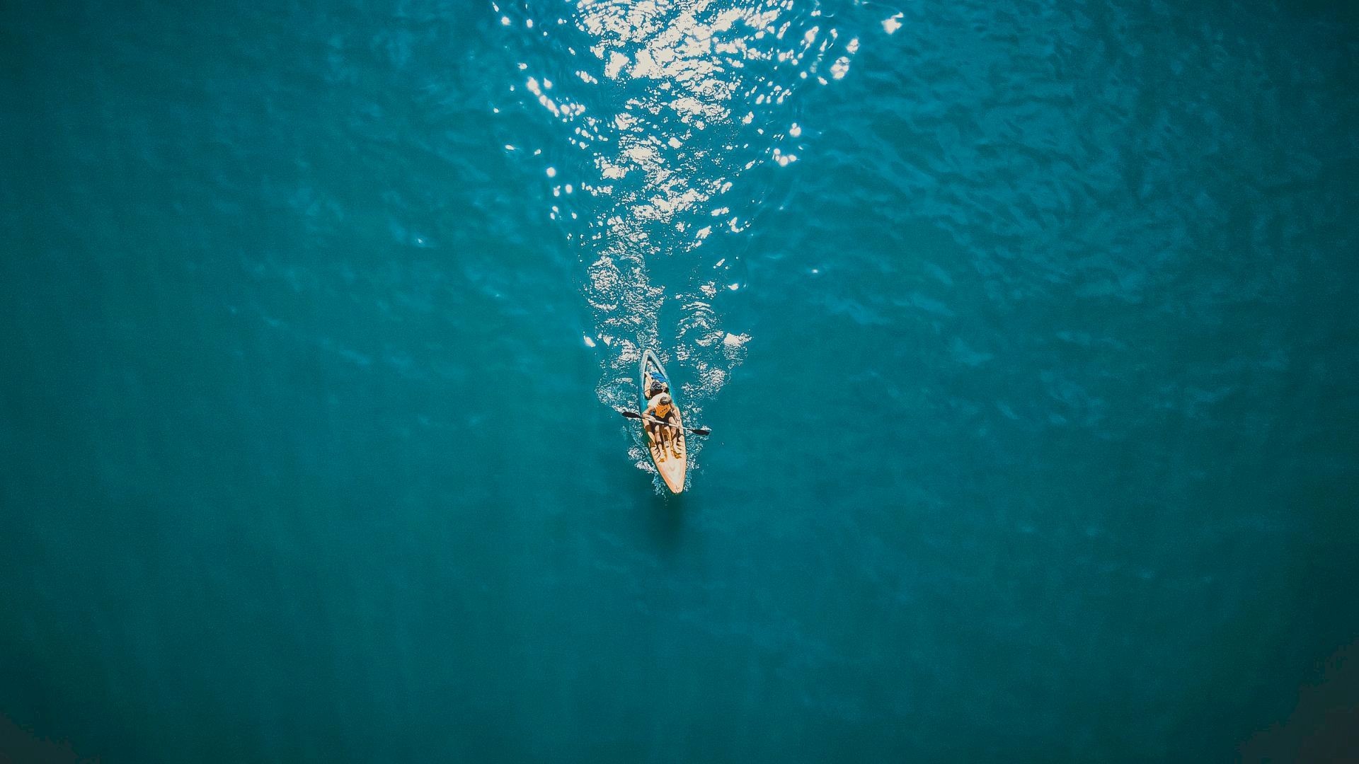 A surfer paddles on a yellow board through clear blue water, leaving a foamy splash behind them.