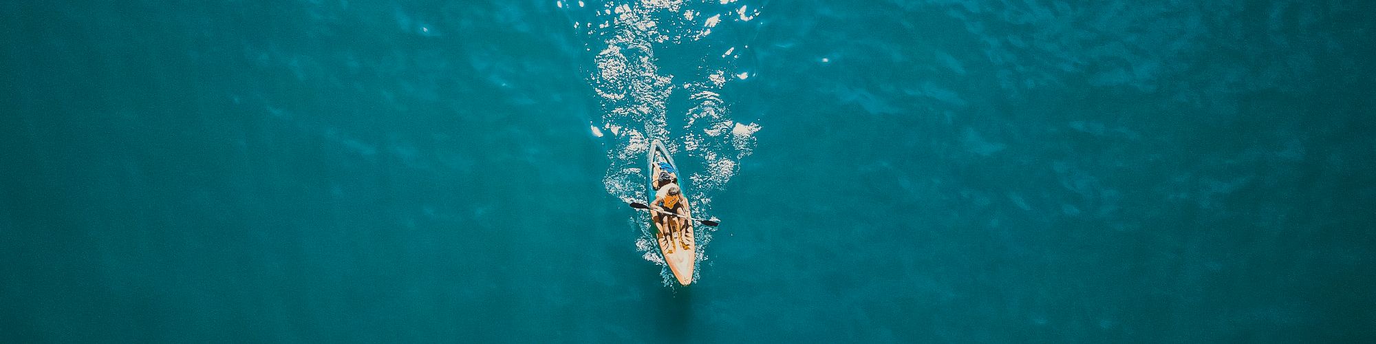 An overhead view of a person kayaking on a vast, calm blue body of water, with the sun reflecting off the surface.