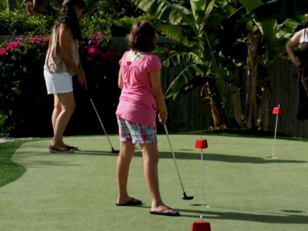 People are playing on a putting green, surrounded by plants and small flags marking holes, in a sunny outdoor setting.