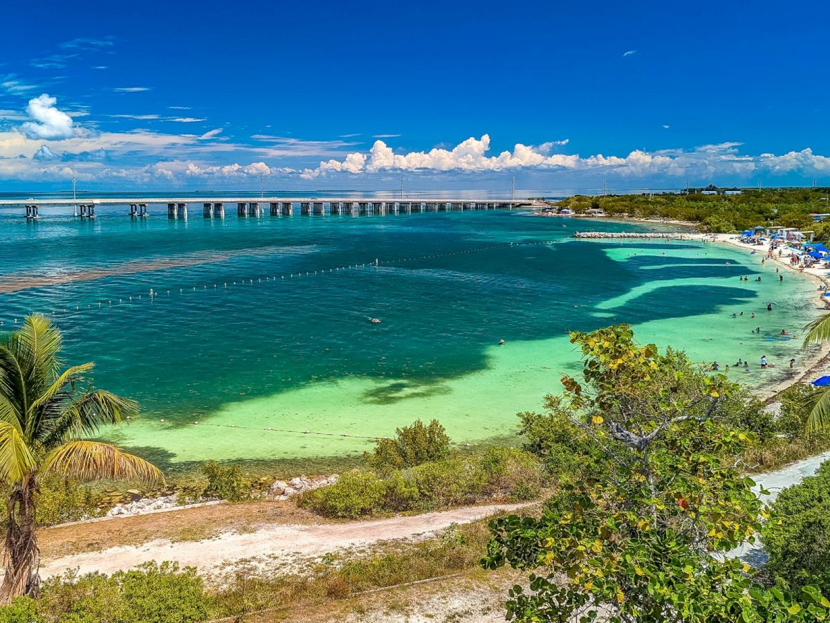 Sunny tropical beach with clear turquoise water and a long pier stretching over the bay under blue skies.