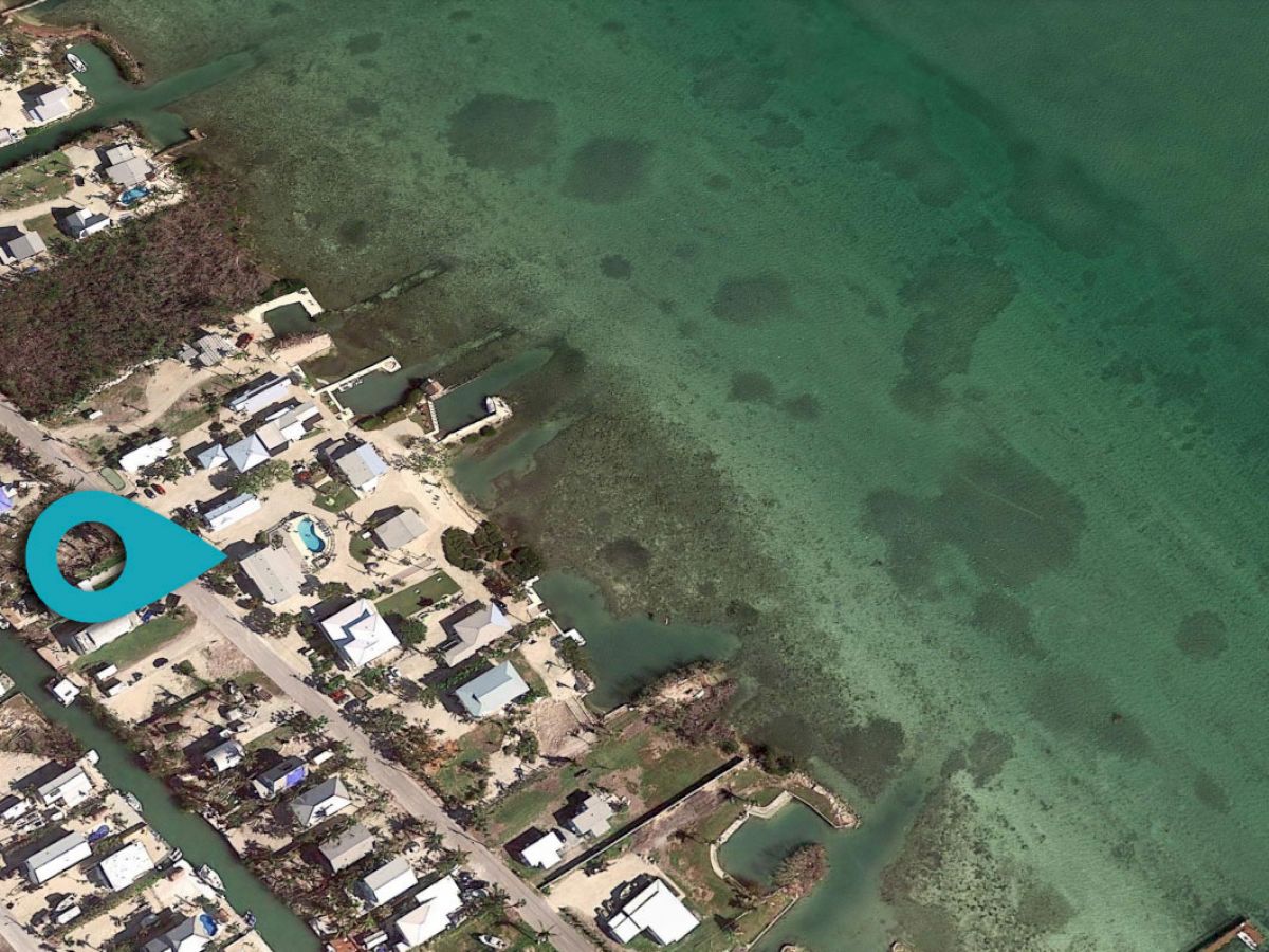 This is an aerial view of a coastal residential area with houses, roads, and docks adjacent to a body of water, marked with a blue pin.