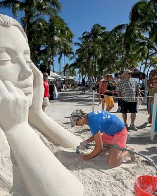 An artist working on a large sand sculpture of a woman's face and hands while people watch on a sunny beach with palm trees and a clear blue sky.