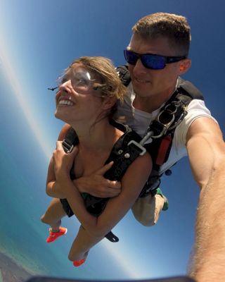 Two people tandem skydiving, with the instructor holding the student. They are free-falling through the sky over a scenic landscape.