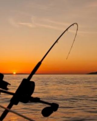 A fishing rod is secured to a boat, silhouetted against a beautiful orange sunset over a calm body of water.