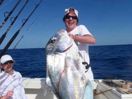 Two people are on a boat in the ocean, with one of them holding a very large fish, smiling. Fishing rods are visible in the background.