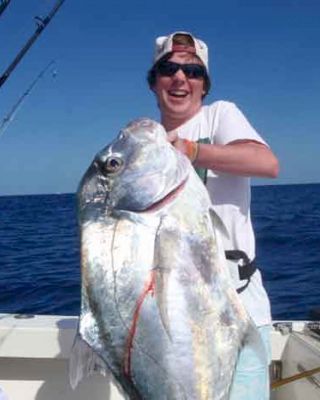 Two people are on a boat in the ocean, with one of them holding a very large fish, smiling. Fishing rods are visible in the background.