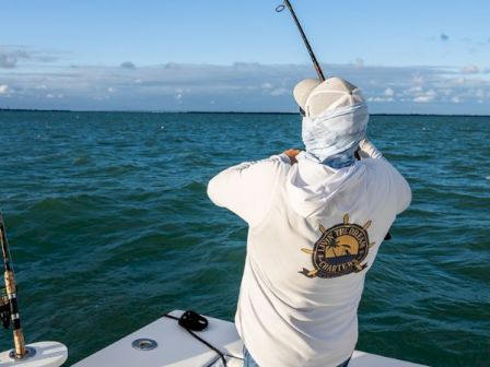 A person wearing a white long-sleeve shirt and head covering fishing from a boat on blue water, with a logo on the back, casting a line.