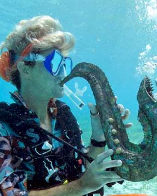 A person is underwater, wearing scuba gear, and playing a saxophone-shaped sculpture that looks like a sea creature, with bubbles rising upwards.