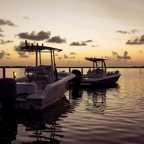 The image features two motorboats tied to a dock on a calm body of water during sunset, with clouds scattered across the sky in the background.