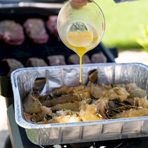 A grill with a foil tray of seasoned seafood as yellow sauce is poured from a measuring cup onto the food.