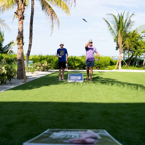 Two men stand on a sunny tropical resort lawn, playing a game on a green court beside palm trees and the ocean.