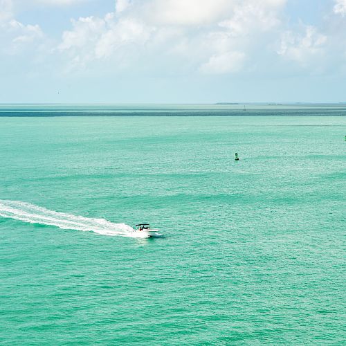 A boat is speeding across a vast expanse of turquoise ocean under a partly cloudy sky, leaving a trail of white wake behind it.