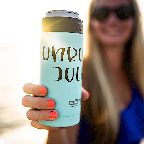 A person is holding a light blue can cooler with "UNRULY JULI" written on it. The background suggests a sunny, outdoor setting by the water.