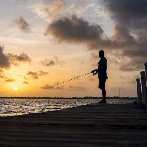 A person is fishing on a dock at sunset, silhouetted against a colorful sky and calm water.