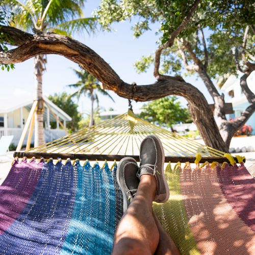 Relaxing in a colorful hammock outdoors under trees, with legs up and sunny sky above.