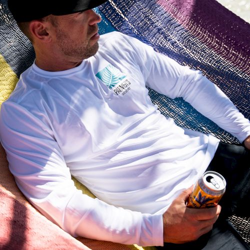 A man in a white resort shirt lounges in a hammock by the water, holding a beer can, soaking up sunny vibes.