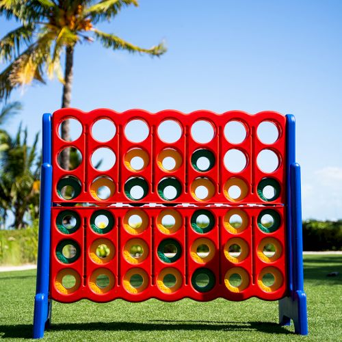 A bright red Connect-style board game with colorful discs stands on green grass beneath swaying palm trees under a clear blue sky.