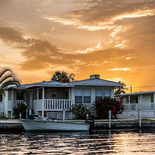 A golden sunset over a waterfront home with palm trees and a small boat docked in front.