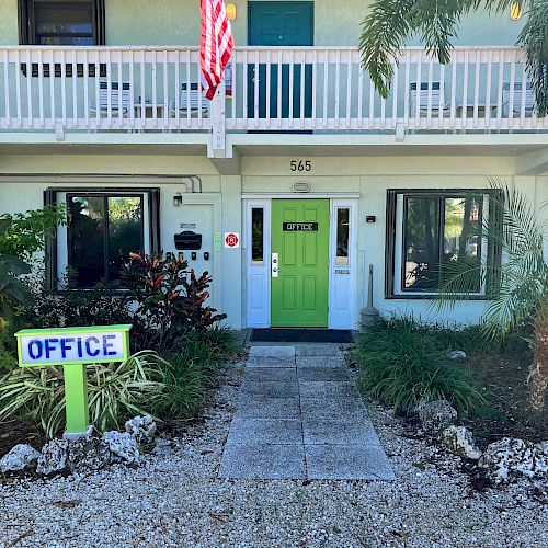 A building with an American flag, green door, "OFFICE" sign, and landscaping. The entrance path is lined with stones and plants, creating a welcoming look.