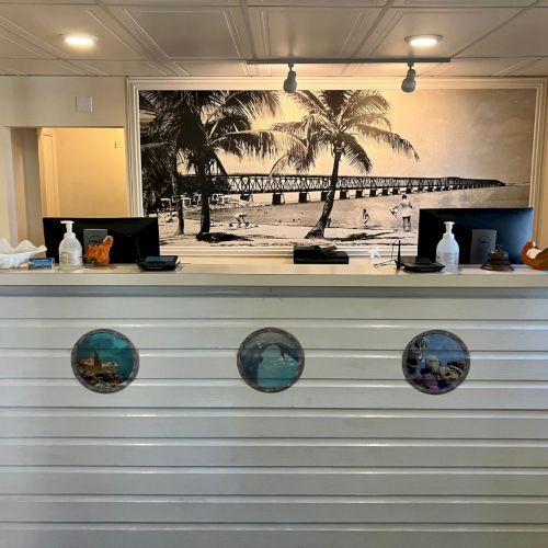 A white reception desk with two computers, sanitizer bottles, and a large palm-tree beach mural backdrop.