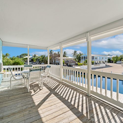A bright seaside veranda with white chairs overlooking a blue pool and lush palms under a sunny sky.