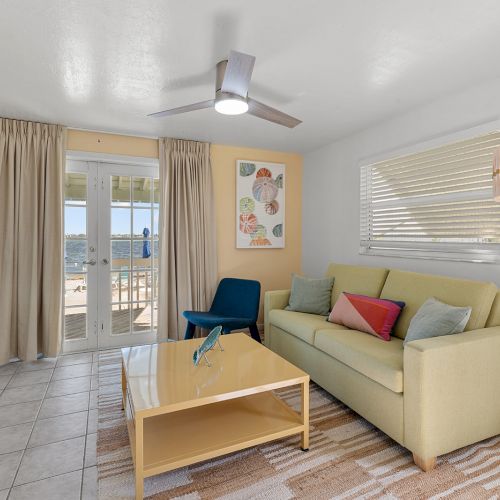 A cozy living room with a sofa, coffee table, wall art, and a TV, leading to a balcony with ocean views.