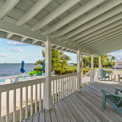 A beachfront porch with Adirondack chairs, overlooking a sandy beach with umbrellas and palm trees by the water, on a sunny day.
