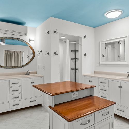 A bright modern bathroom with two white vanities, double sinks, a wood countertop, and large mirrors above a tiled room.
