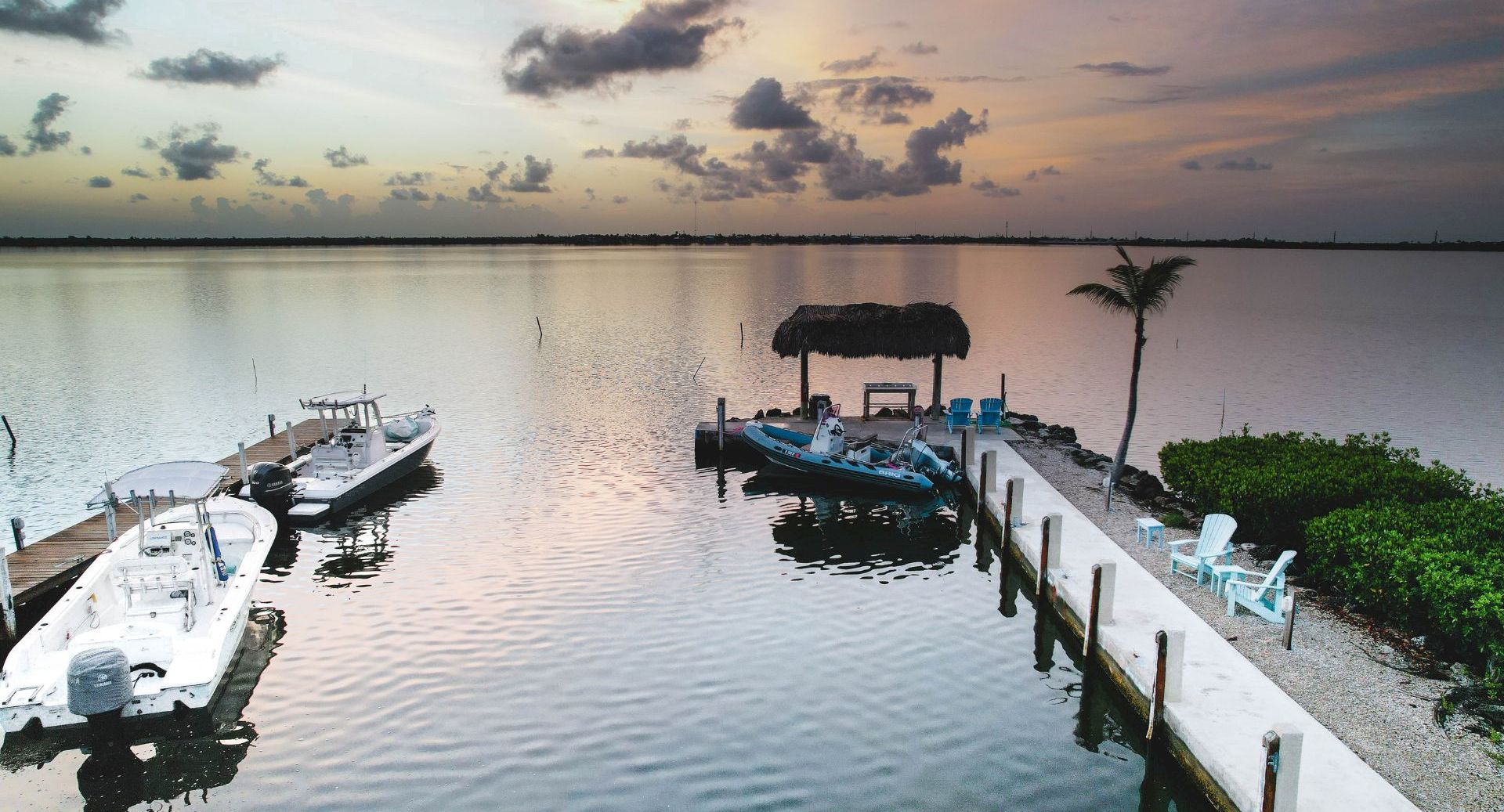 Boats docked at a quiet marina by calm water at sunset, with a palm tree and thatched shade in the foreground.