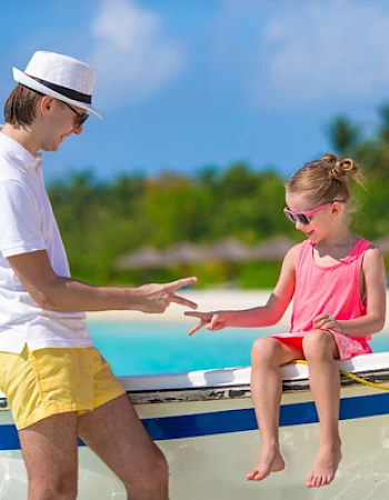 A man and a child are playing rock-paper-scissors on a boat by a tropical beach, both wearing summer outfits and sunglasses.