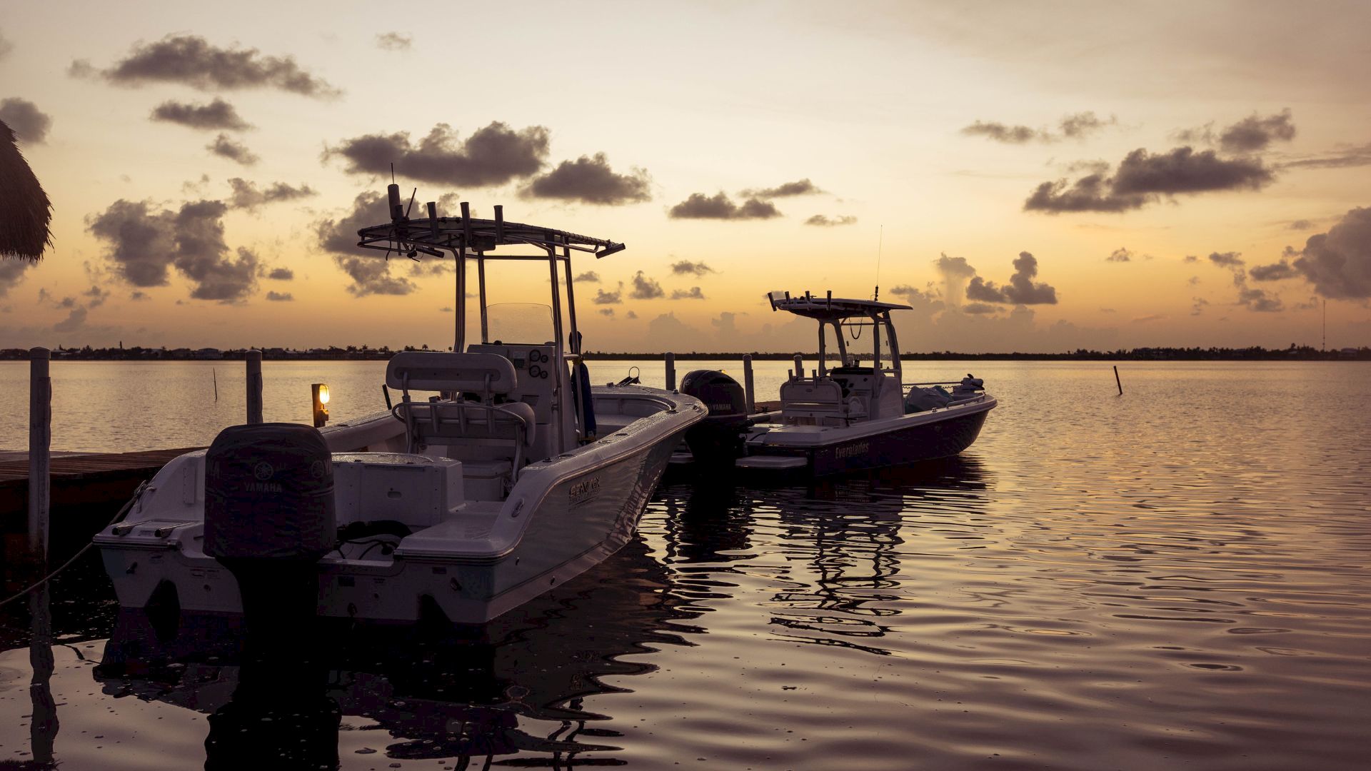 A motorboat with outboard engines is moored at a dock as the sun sets over calm water.
