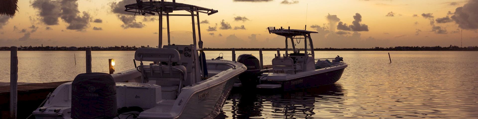 A motorboat with outboard engines is moored at a dock as the sun sets over calm water.