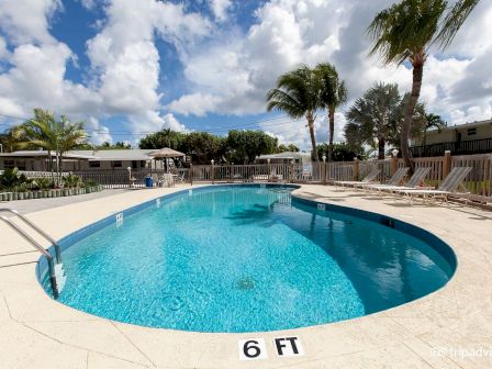 A clear outdoor swimming pool, surrounded by lounge chairs and palm trees under a partly cloudy sky, with "6 FT" marked at the pool’s edge.