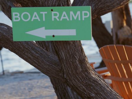 Sign points to a BOAT RAMP; green sign on a tree with a left arrow, beach/ocean in background.