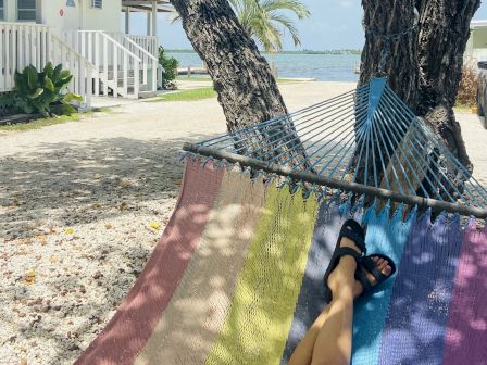A person relaxes on a colorful hammock under a tree by a beach, with cabins, palm trees, and the sea in the background.