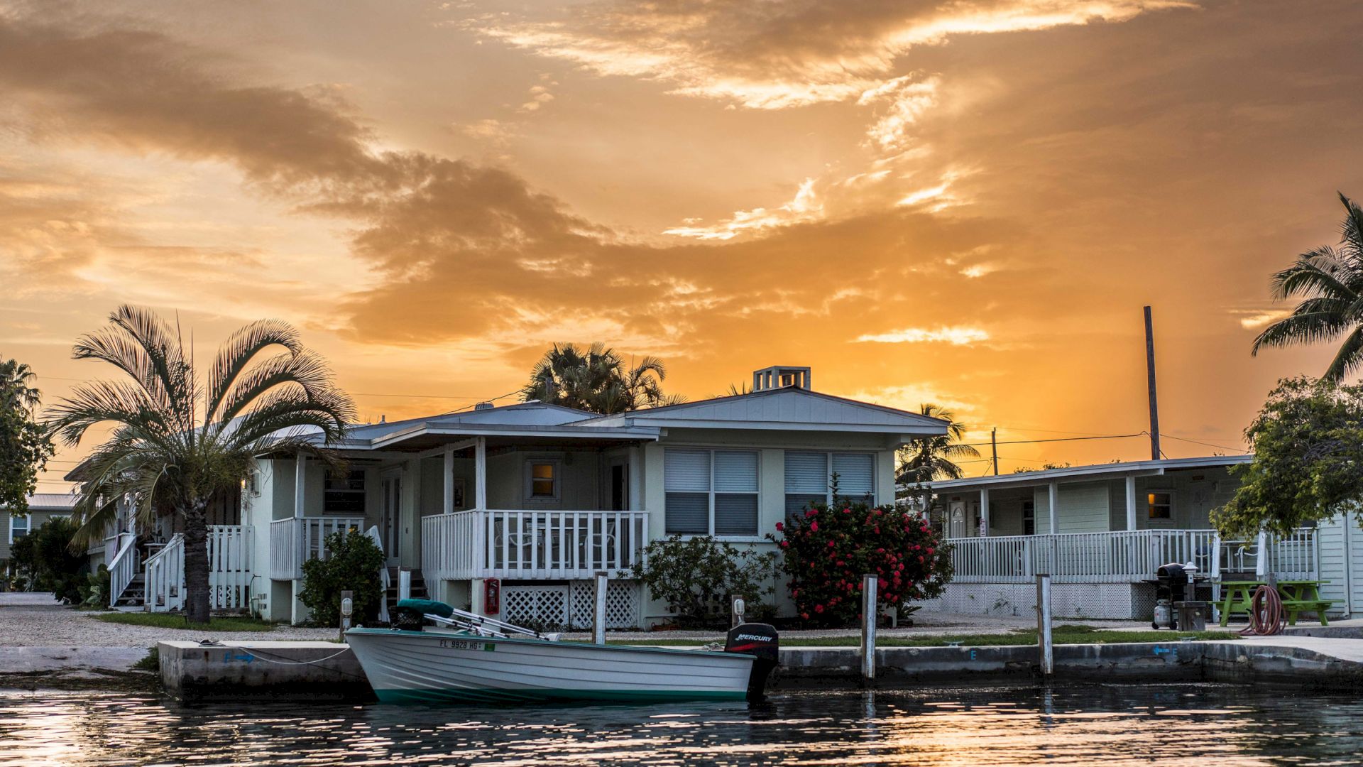 A serene waterfront scene depicts a cozy house with a boat docked nearby, framed by palm trees and bathed in a warm, glowing sunset.