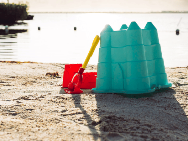 A blue sandcastle mold, a yellow shovel, and a red bucket on a sandy beach near calm water with a shoreline and distant background.