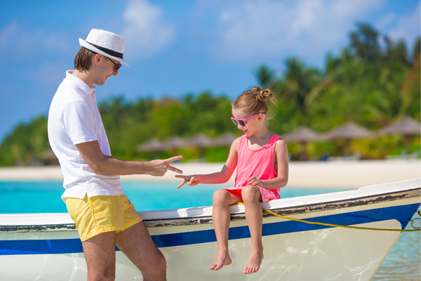 A man and a child are playing rock-paper-scissors on a boat by a tropical beach, with palm trees and clear skies in the background.