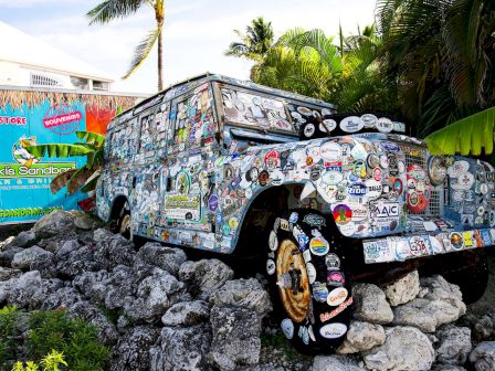 A sticker-covered off-road pickup truck sits on rocky ground near palm trees, surrounded by colorful souvenir signs.