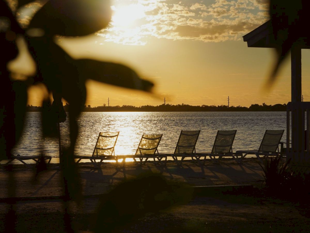 Sunset over a calm lake with lounge chairs in the foreground and a silhouetted person, framed by dock rails.