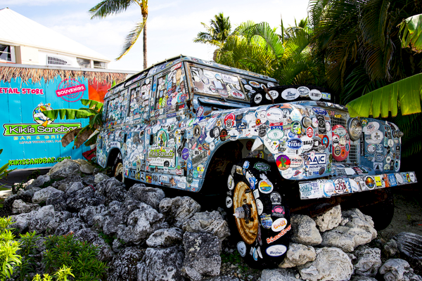 A vehicle covered in stickers is mounted on rocks near palm trees and a colorful sign in a tropical setting.
