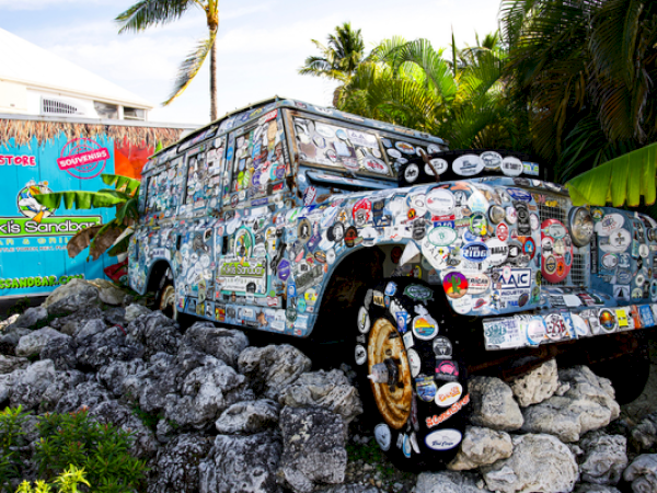 A vehicle covered in stickers is mounted on rocks near palm trees and a colorful sign in a tropical setting.