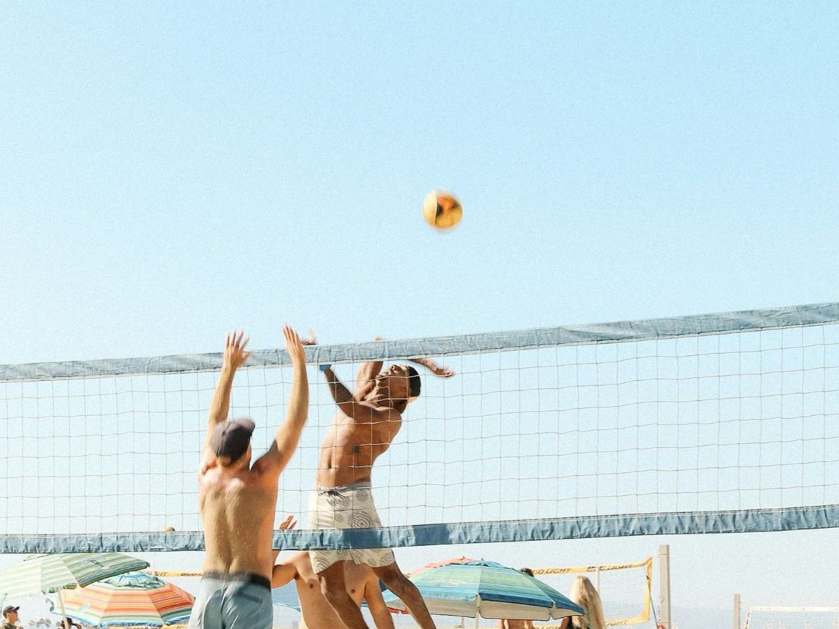 People play volleyball on a sunny beach, with a ball arcing above the net against a bright sky.
