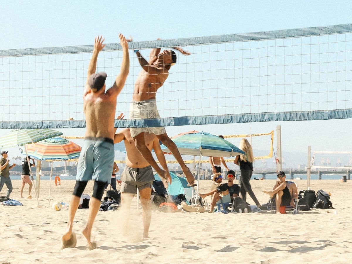 People are playing beach volleyball on a sandy beach with others watching, and a clear blue sky above.