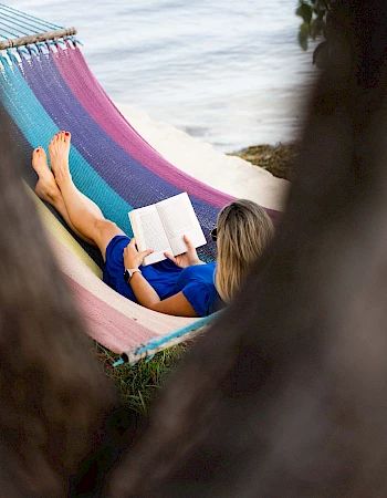 A person relaxing in a colorful hammock by the beach, reading a book, framed by tree branches. Top it at 140 characters, always ending the sentence.